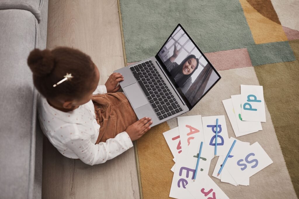A young girl participates in an online learning session with alphabet cards and a laptop.