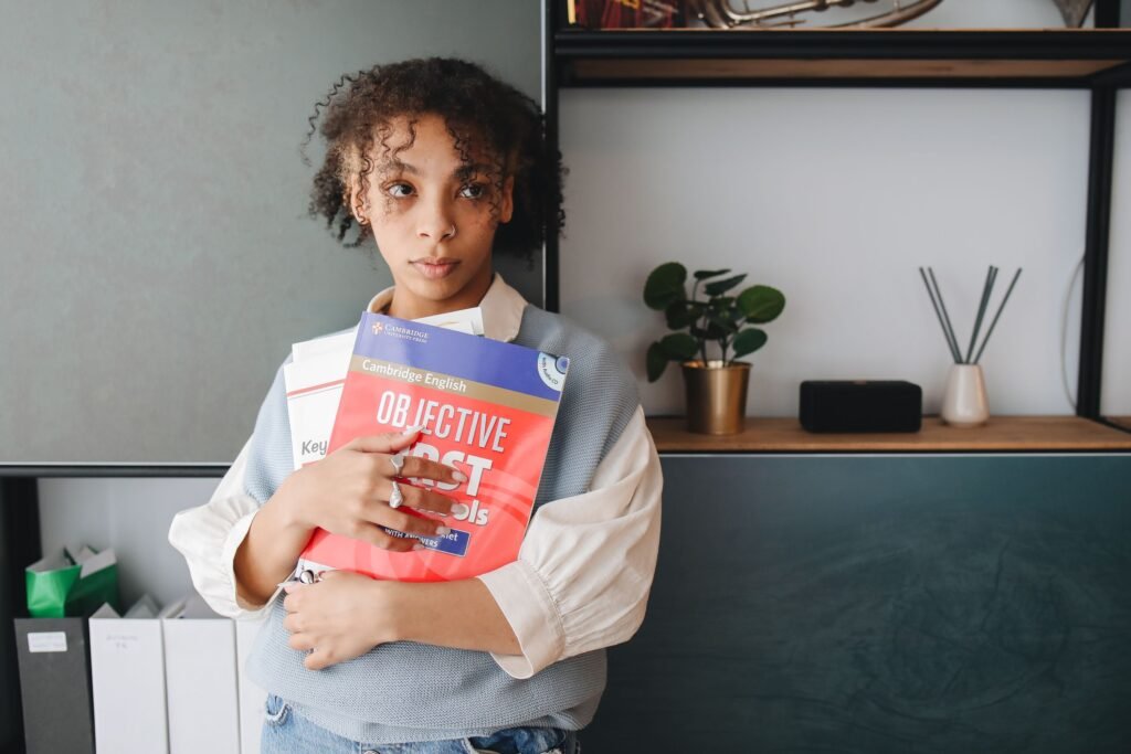 A young woman holding Cambridge English books, standing indoors with a thoughtful expression.